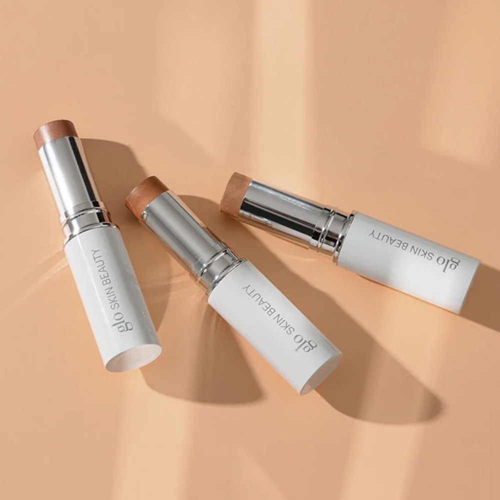 Glo Skin Beauty Skin Glow Stick Highlighter Glo Skin Beauty Skin Glow Stick Highlighter - trzy kremowe rozświetlacze w sztyfcie w odcieniach Champagne, Rosé i Cognac, eleganckie biało-srebrne opakowania na brzoskwiniowym tle, efekt naturalnego blasku i rozświetlenia skóry.