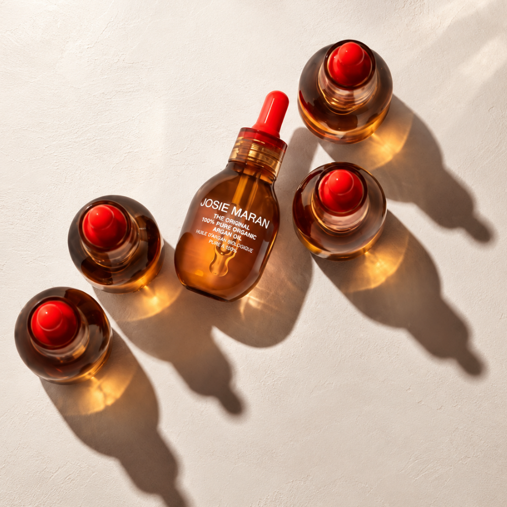 Josie Maran Pure Organic Argan Oil (2) Pięć bursztynowych buteleczek olejku Josie Maran The Original 100% Pure Organic Argan Oil z czerwonymi pipetami, ułożonych na jasnym, beżowym tle. Promienie słońca podkreślają złocisty kolor olejku arganowego, tworząc ciepłe refleksy i wyraziste cienie.