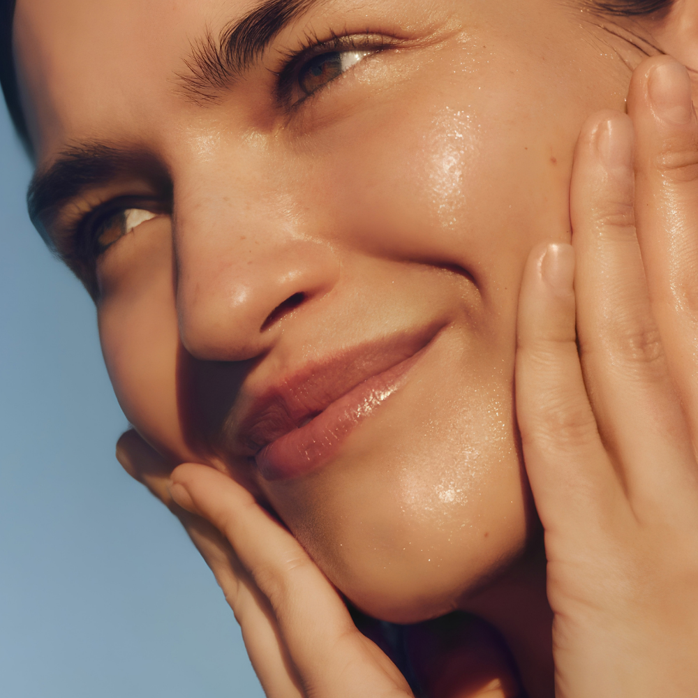 Josie Maran Pure Organic Argan Oil (4) Uśmiechnięta modelka o promiennej, rozświetlonej cerze delikatnie dotyka twarzy dłońmi, prezentując efekt pielęgnacji Josie Maran The Original 100% Pure Organic Argan Oil - organicznym olejkiem arganowym do twarzy, który pomaga odżywiać, wygładzać i zmiękczać skórę.