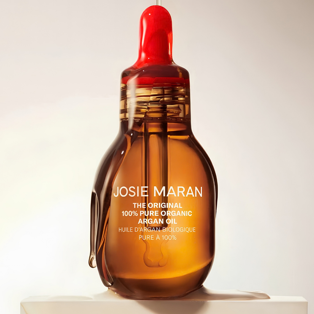 Josie Maran Pure Organic Argan Oil Szklana buteleczka Josie Maran The Original 100% Pure Organic Argan Oil z czerwonym zakraplaczem, zawierająca czysty organiczny olejek arganowy do pielęgnacji twarzy, ciała i włosów, na jasnym beżowym tle.