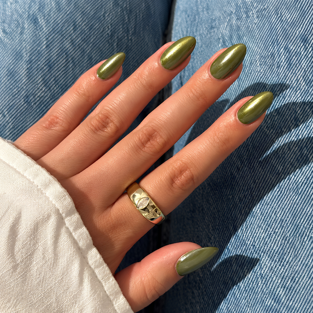 Lights Lacquer Glazed Pearl Powder Stick (4) Manicure w odcieniu oliwkowej zieleni z metalicznym, perłowym połyskiem i efektem glazed nails. Długie paznokcie w kształcie migdała ozdobione błyszczącym wykończeniem, dłoń ze złotym pierścionkiem na tle niebieskiego jeansu.