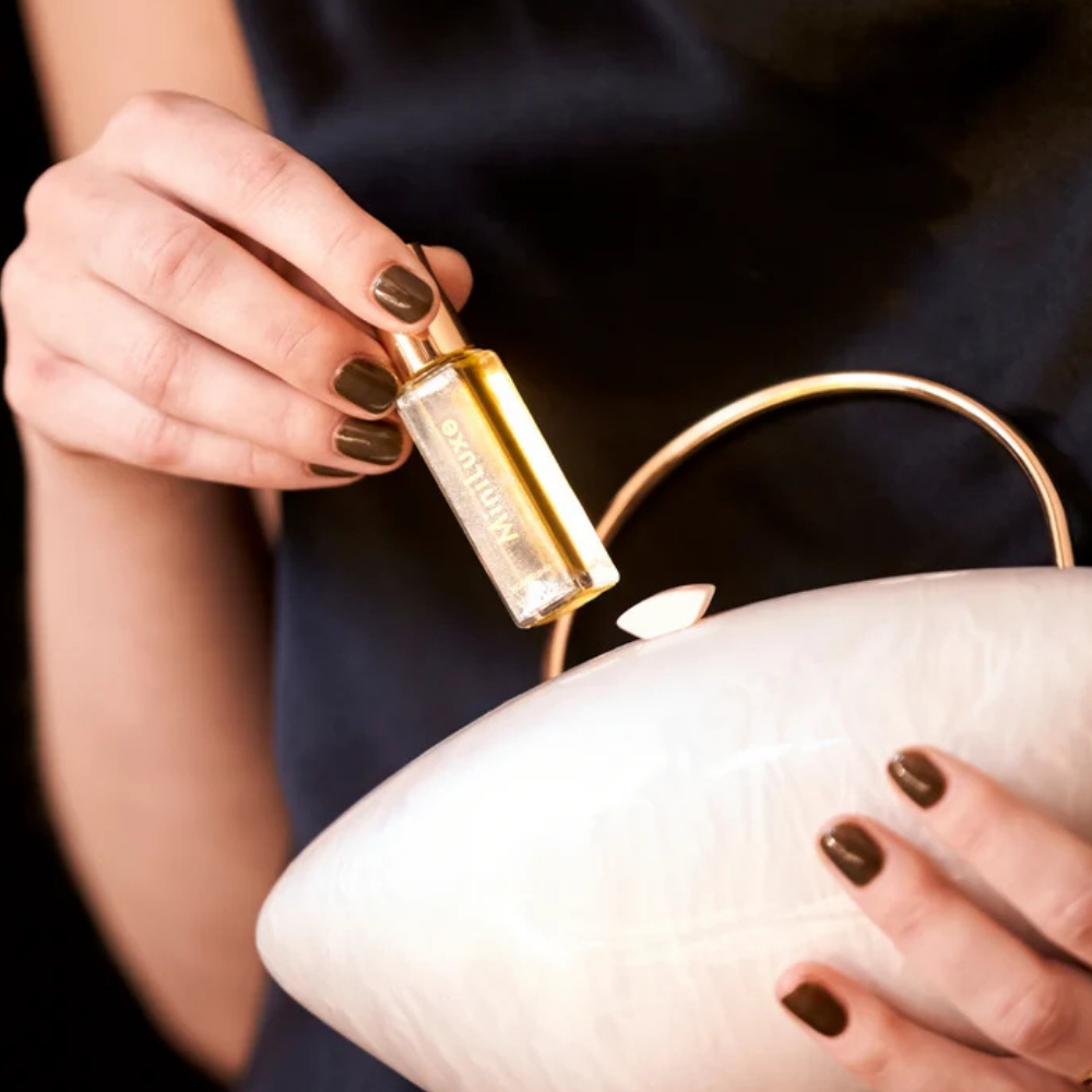 Mini Luxe Nourishing Cuticle Oil (4) Kobieta z ciemnobrązowym manicure trzyma odżywczą oliwkę MiniLuxe Nourishing Oil nad jasną torebką. Elegancka fotografia lifestyle podkreślająca poręczny rozmiar produktu.