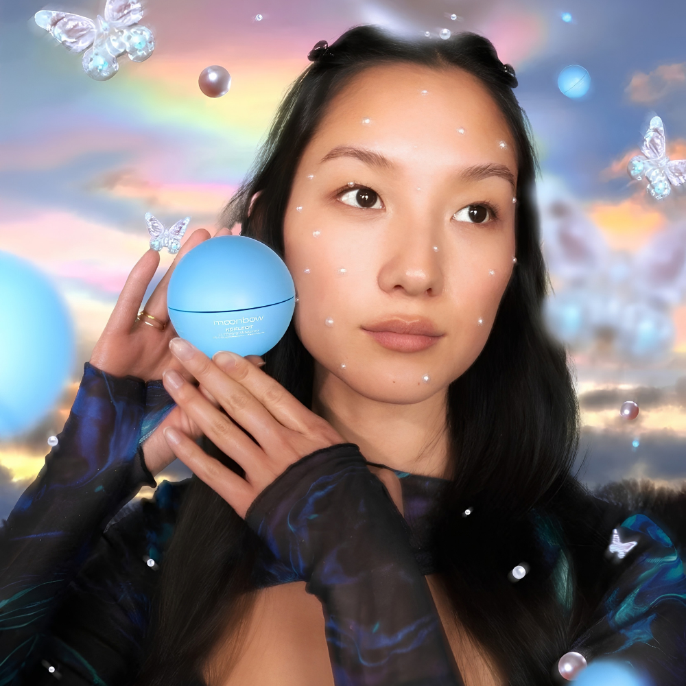 Moonbow Reflect Luminizing Moisturizer (2) Modelka trzyma niebieskie opakowanie Moonbow Reflect Luminizing Moisturizer przy twarzy z nałożonym kremem, na pastelowym tle z perłami i motylami.