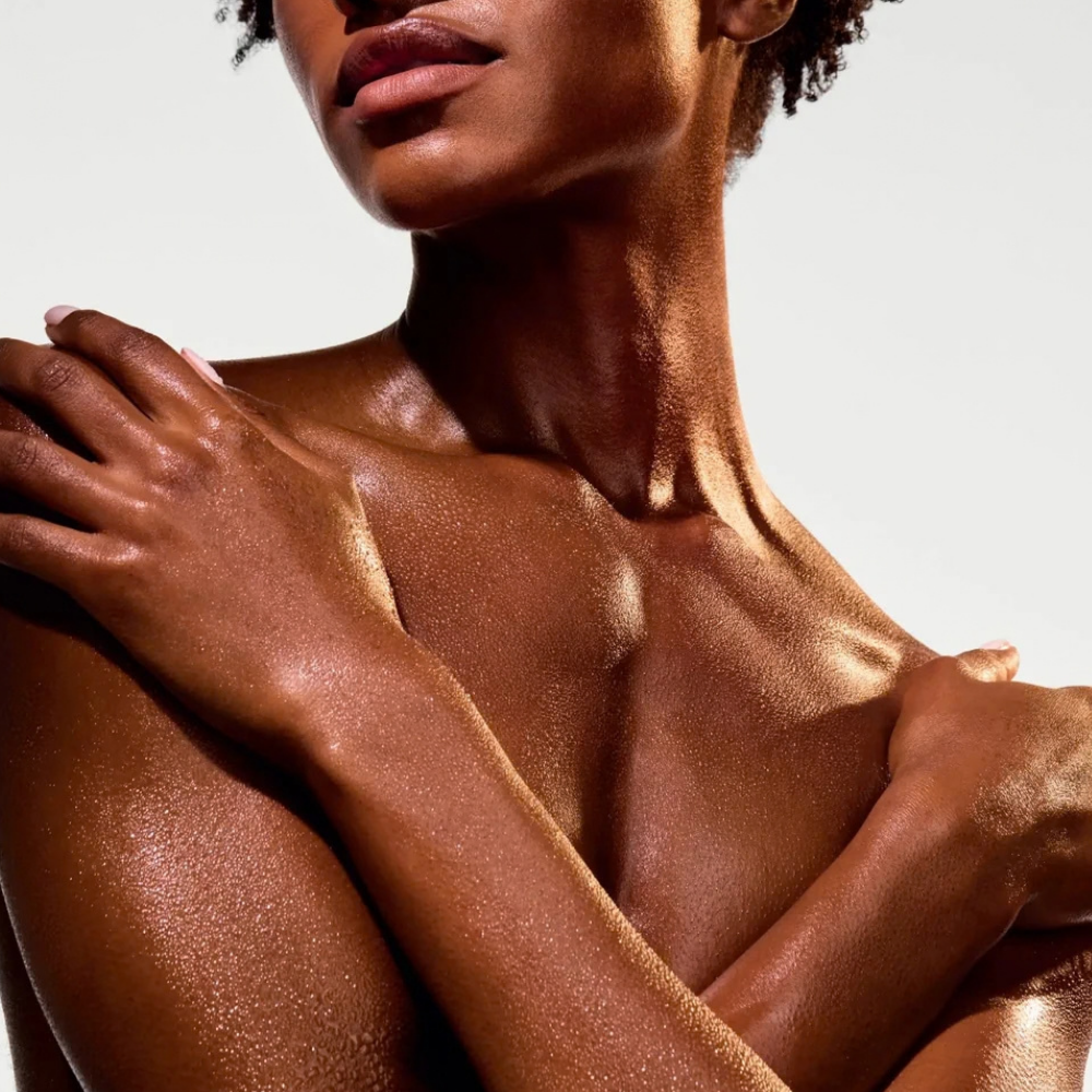 Naturium The Glow Getter Multi Oil Body Scrub (6) Naturium The Glow Getter Multi Oil Body Scrub - zbliżenie na modelkę o ciemnej karnacji, z ramionami skrzyżowanymi na dekolcie. Jej gładka, nawilżona skóra pięknie odbija światło, podkreślając jedwabiste wykończenie i naturalny blask po pielęgnacji ciała.