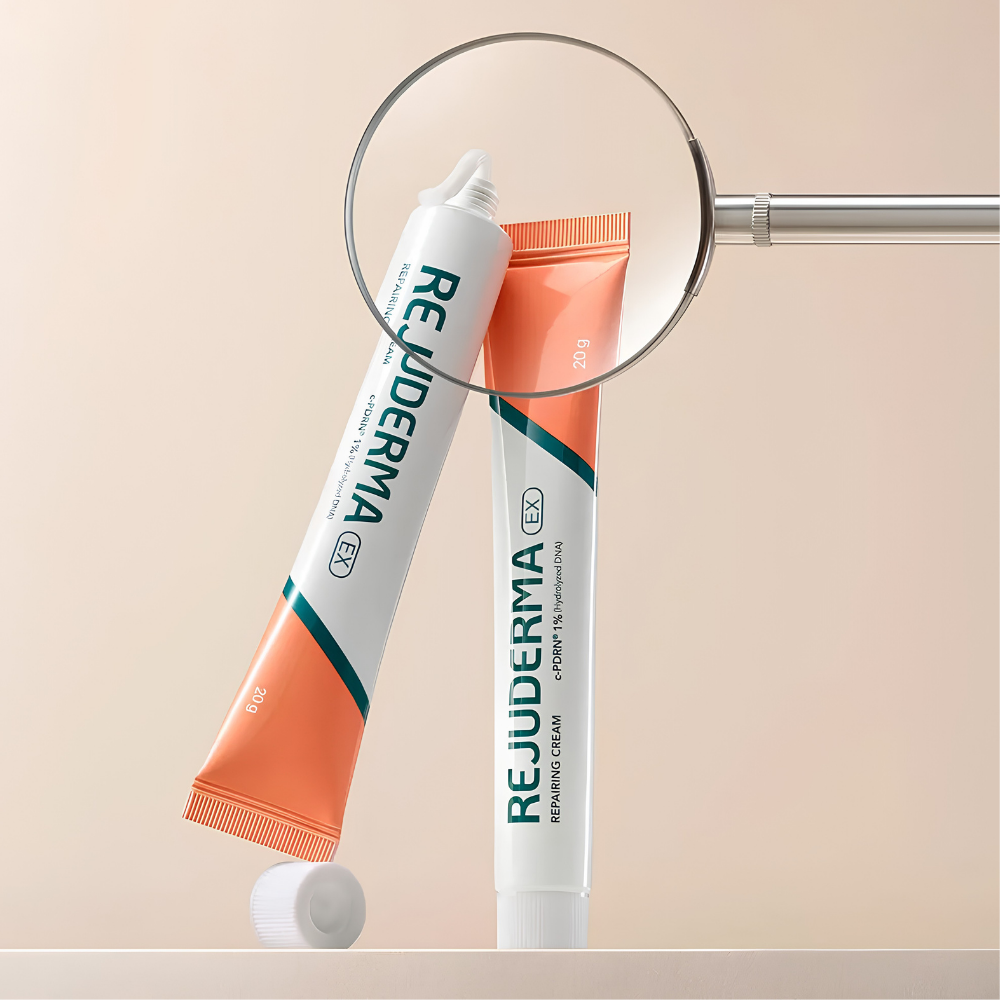 Rejuderma Ex Repairing Cream c PDRN 1% REJUDERMA EX Repairing Cream c-PDRN 1% - koreański krem kojąco-odbudowujący do twarzy, dwie biało-koralowe tubki 20 g z zielonym logo marki, jedna otwarta z widoczną konsystencją kremu, elegancka kompozycja z lupą na beżowym tle.