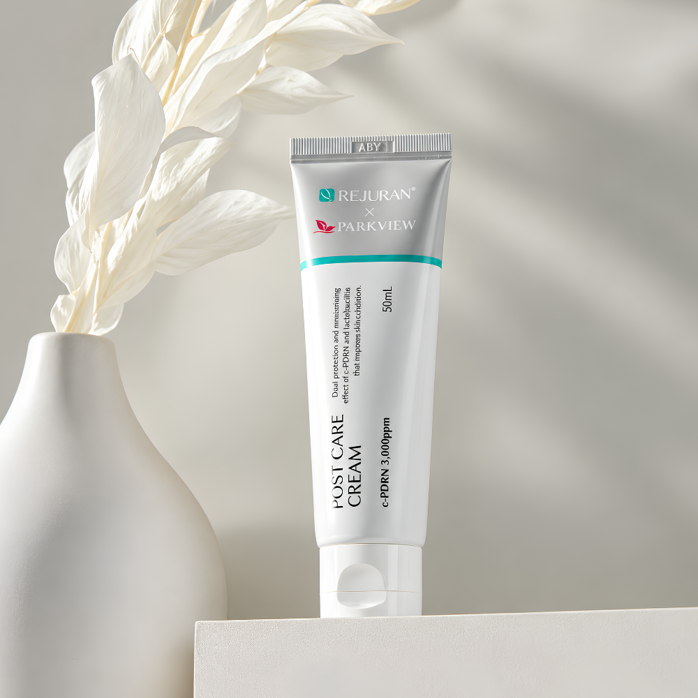 Rejuran X Parkview Post Care Cream (3) REJURAN x PARKVIEW Post Care Cream c-PDRN 3000 ppm - Koreański krem regenerujący i kojący do twarzy, 50 ml, w srebrno-białej tubce z turkusowym akcentem, ustawiony na jasnym podeście obok białego wazonu z dekoracyjnymi liśćmi.