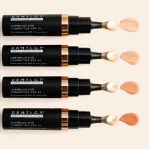 Na zdjęciu widoczne są cztery tubki produktu o nazwie "DRMTLGY MEDICAL GRADE SKIN CARE LUMINOUS EYE CORRECTOR SPF 41 Broad Spectrum SPF 41 Sunscreen". Każda tubka ma aplikator z próbką produktu obok, co wskazuje, że korektor pod oczy jest dostępny w różnych odcieniach, aby dopasować się do różnych tonów skóry. Tubki są czarne z akcentami w kolorze złotym i białym, co nadaje im luksusowy wygląd. Produkt do pielęgnacji skóry wokół oczu z dodatkową ochroną przeciwsłoneczną.