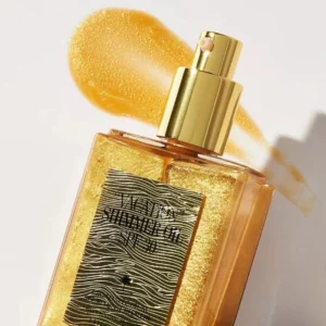 Na obrazku znajduje się elegancka butelka złotego olejku "Vacation Shimmer Oil SPF 30" z atomizerem, który ma złoty dozownik. Butelka jest częściowo przechylona, a w tle widoczny jest złocisty ślad po aplikacji olejku, który wygląda na kremowy i błyszczący. Etykieta na butelce ma wzór przypominający fale lub teksturę drewna, co dodaje produktowi wyjątkowego charakteru. Olejek wygląda na luksusowy i ma intensywnie złoty kolor, który sugeruje, że dodaje skórze połyskującego blasku. Całość jest przedstawiona na jasnym tle, co podkreśla złocisty kolor olejku.