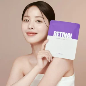 Uśmiechnięta kobieta o promiennej cerze trzyma opakowanie Lapcos Retinal Resurfacing Face Mask, prezentując je na tle ciepłego beżowego odcienia. Opakowanie w eleganckiej biało-fioletowej kolorystyce podkreśla kluczowy składnik – retinal, znany ze swoich właściwości wygładzających i odnawiających skórę. Jej świeży i zdrowy wygląd sugeruje nawilżające i odżywcze działanie produktu.