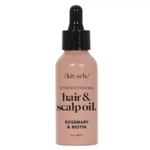 Hair 13 Różowa butelka z czarną zakrętką i pipetą. Na butelce znajduje się napis: "/kit·sch/ STRENGTHENING hair & scalp oil. ROSEMARY & BIOTIN 2 oz. (60mL)". Produkt przeznaczony jest do wzmacniania włosów i skóry głowy, zawiera rozmaryn i biotynę.