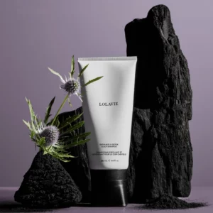 Hair 4 Szampon Lolavie „Exfoliate & Detox Scalp Shampoo” jest ustawiony na tle ciemnych skał, obok dekoracji w postaci kwiatów i roślin. Kompozycja zdjęcia podkreśla naturalne składniki oraz oczyszczające właściwości produktu, a fioletowe tło dodaje elegancji.
