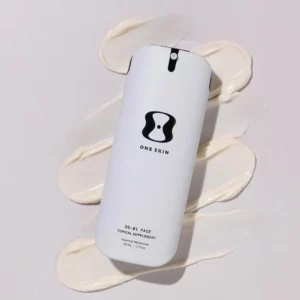 Start 130 Biały flakon z minimalistycznym designem marki One Skin, na którym widnieje logo w kształcie klepsydry i napisy: "OS-01 Face Topical Supplement" oraz "Essential Moisturizer, 50 mL / 1.7 fl oz". Produkt leży na tle rozsmarowanego kremu o gładkiej, kremowej konsystencji, tworzącego estetyczną kompozycję.