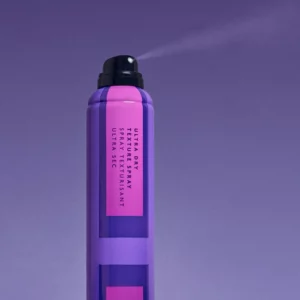 Na zdjęciu znajduje się fioletowy spray "Ultra Dry Texture Spray". Produkt jest aplikowany przez dyszę umieszczoną na szczycie butelki, a na tle widać drobną mgiełkę kosmetyku, co wskazuje na suchą, lekką konsystencję rozpylaną podczas aplikacji.