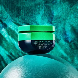 Na zdjęciu znajduje się słoiczek serum do włosów na noc R+Co Bleu Sleep Masque Night Repair. Opakowanie ma jasnozieloną zakrętkę. Produkt umieszczony jest na wypukłej, zielonej powierzchni, a tło tworzy teksturowana ściana w odcieniach zieleni i niebieskiego.