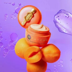 Otwarty słoiczek Tatcha The Brightening Eye Cream o intensywnie pomarańczowym kolorze, wypełniony kremem o jasnej, kremowej konsystencji, ustawiony na tle moreli i fioletowej, przezroczystej tekstury symbolizującej nawilżenie.