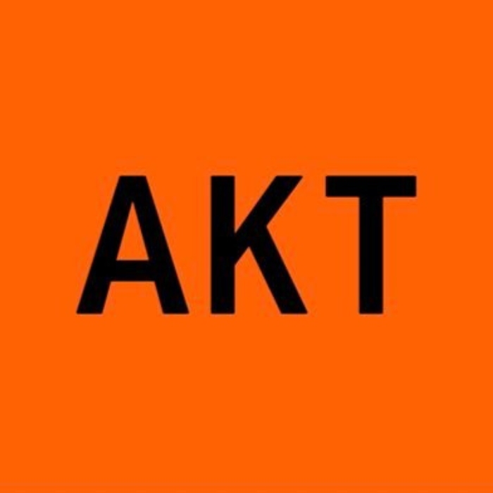 Na zdjęciu znajduje się logo marki AKT w czarnym kolorze, na intensywnie pomarańczowym tle, które podkreśla minimalistyczny design.