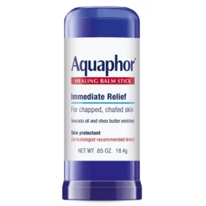 Zdjęcie przedstawia sztyft ochronny do skóry Aquaphor Healing Balm Stick w plastikowym opakowaniu o niebieskim kolorze. Na etykiecie znajduje się nazwa marki "Aquaphor" oraz informacja, że produkt zapewnia natychmiastową ulgę dla spierzchniętej i podrażnionej skóry. Zawiera olej z awokado i masło shea, które odżywiają i nawilżają skórę. Produkt jest oznaczony jako ochrona skóry i rekomendowany przez dermatologów. W dolnej części etykiety widnieje informacja o pojemności: (18,4 g).