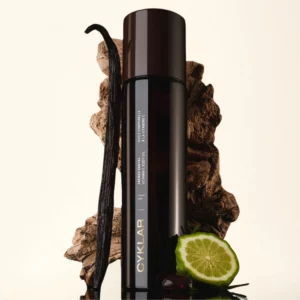 Cyklar Sacred Santal Vitamin C Body Oil ustawiony przed ciemnym tłem z drewnem, waniliową laską i plasterkiem bergamotki, podkreślającym zmysłowy i cytrusowo-drzewny charakter olejku.