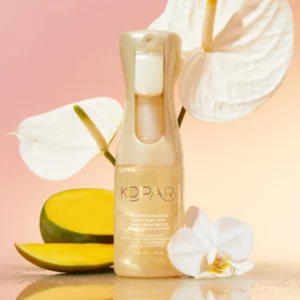 Kopari Golden Sunglaze Sheer Body Mist SPF 50 na tle kwiatów storczyka i plastra mango, w pastelowym, wakacyjnym klimacie.
