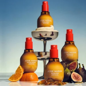 Cztery butelki mgiełek zapachowych Josie Maran ustawione na lustrzanym tle: „Vanilla Vibezzz” na szczycie kielicha z dekoracyjną bitą śmietaną, „Topless Tangerine” oparta na pomarańczy z plasterkiem owocu, „Golden Hour” w otoczeniu bursztynów oraz „Bohemian Fig” przy świeżych figach. Tło w odcieniach błękitu podkreśla letni, zmysłowy klimat kolekcji.