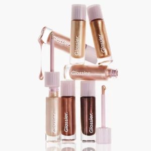 Zestaw płynnych cieni do powiek Glossier Skylight w różnych odcieniach – od perłowej bieli, przez złoto, brąz, po miedziane i szampańskie tony. Na zdjęciu widoczne są buteleczki z aplikatorami oraz kropla produktu, podkreślająca ich kremową konsystencję i połyskujące wykończenie.