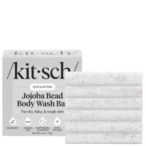 Białe opakowanie kostki Kitsch Jojoba Bead Body Wash Bar wraz z samą kostką o żłobionej powierzchni i widocznymi czarnymi drobinkami jojoba.