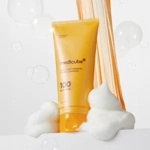 Sale 7 Żółta tubka produktu do oczyszczania twarzy Medicube Kojic Acid Turmeric Toning Cleanser ustawiona pionowo na białym tle, otoczona puszystą pianą i przezroczystymi mydlanymi bąbelkami. W tle widoczna złocisto-pomarańczowa smuga nawiązująca do koloru kurkumy.
