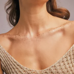 Kobieta w złotej, siateczkowej sukni z dekoltem ozdobionym subtelnymi złotymi drobinkami Fazit Decolletage Speckles Gold, które dodają skórze blasku i elegancji.