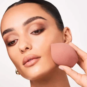 Kobieta przykłada gąbkę Patrick Ta Skin 5 In 1 Blending Sponge do policzka, demonstrując aplikację makijażu twarzy.
