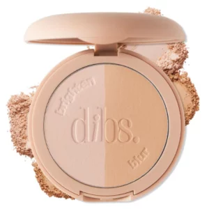 Otwarty puder DIBS LoveBeam Setting Powder w beżowym opakowaniu, podzielony na dwie części: jaśniejszą „brighten” i ciemniejszą „blur”. Wokół widoczne rozsypane drobinki pudru w odcieniach jasnego beżu, na białym tle.