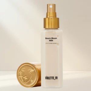 Butelka z atomizerem Boum-Boum Milk (3-in-1 cream spray) marki Violette_FR. Mleczna formuła widoczna w przezroczystym opakowaniu ze złotą pompką; obok leży złota nakrętka z wytłoczonym logo. Produkt ustawiony na jasnym, ciepłym tle podkreślającym minimalistyczny, elegancki charakter.