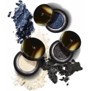 Cienie do powiek Victoria Beckham Lid Lustre – otwarte słoiczki z intensywnie połyskującymi pigmentami w odcieniach granatowym, szampańskim i grafitowym, otoczone rozsypanym produktem na białym tle.