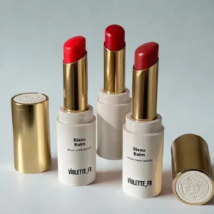 Zbliżenie na trzy pomadki Bisou Balm (sheer matte lipstick) marki Violette_FR w białych, minimalistycznych opakowaniach ze złotymi elementami. Sztyfty w odcieniach czerwieni i malinowego różu są wysunięte, obok leżą złote zatyczki z wytłoczonym logo. Jasne, neutralne tło podkreśla elegancki, francuski charakter produktu.