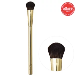 Pędzel do makijażu Sarah Creal The Complexion Brush z pozłacaną rączką i miękkim włosiem na białym tle.