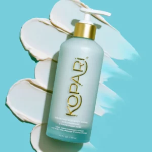 Błękitna butelka z pompką Kopari Hydrating Silk Body Cream leży na rozsmarowanej warstwie kremu, na błękitnym tle; złote detale na opakowaniu podkreślają elegancję produktu.