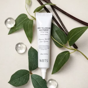 Nōts Centella Scar Ointment – punktowa maść pielęgnacyjna z wąkrotką azjatycką w białej tubce, ułożona na jasnym tle z zielonymi liśćmi roślin i dekoracyjnymi szklanymi kulkami.
