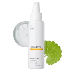 Biała butelka mgiełki Troxederm Repair Essence Mist na jasnym tle z kroplą żelu i liściem wąkroty azjatyckiej w tle.