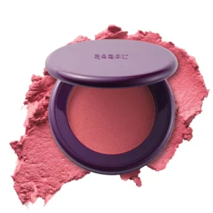 Róż do policzków Sarah Creal Coming in Hot Blurring Pressed-Mousse Blush w odcieniu Love Bites w opakowaniu na tle rozproszonego pigmentu.
