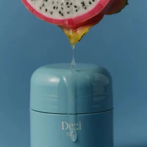 Travel 11 Niebieski słoiczek Dezi Skin Agua Fresca z kroplą soku spływającą z przekrojonej pitai na wieczko, na niebieskim tle.