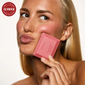 Kobieta prezentująca Glossier Cloud Paint Plush Blush przy policzku, z widocznym różowym rumieńcem i opakowaniem produktu.