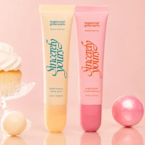 Dwa błyszczyki-balsamy Sincerely Yours Sugarcoat Gloss Balm w odcieniach Sheer Vanilla i Bubbleberry ustawione obok siebie na pastelowym różowym tle. Wokół produktów znajdują się dekoracyjne kulki i waniliowy deser podkreślające cukierkowy charakter kolekcji.