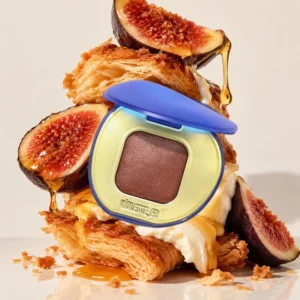 Otwarty Violette_FR Plume Eyeshadow w brązowo-śliwkowym odcieniu, ustawiony w niebiesko-złotym opakowaniu w aranżacji z figami, miodem, kremem i chrupiącym ciastem.