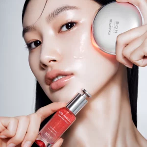 Modelka przykłada urządzenie BOH Bioheal Shurinka do policzka i trzyma czerwone serum NAD+ Prizcell Glow Power Serum.
