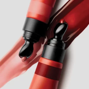 Dwa błyszczyki Hung Vanngo Glossy Lip Shine w koralowo-czerwonych opakowaniach, pokazane na tle smug błyszczącej, różowej i głębokiej czerwonej formuły. Czarne aplikatory pokryte są połyskującym błyszczykiem.