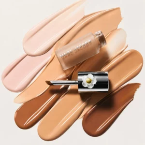 Na zdjęciu znajduje się Marc Jacobs Beauty Nude Awakening Concealer z aplikatorem na tle kremowych swatchy w różnych odcieniach beżu.