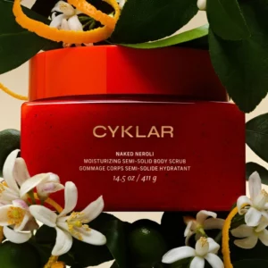 Start 54 Cyklar Naked Neroli Scrub - nawilżający peeling do ciała w pomarańczowo-czerwonym opakowaniu, otoczony kwiatami neroli, zielonymi liśćmi i skórką pomarańczy.