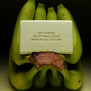 Start 53 Soft Services Green Banana Buffing Bar Microcrystal Exfoliant - zielona kostka peelingująca do ciała zaprezentowana na tle niedojrzałych zielonych bananów.