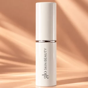 Start 55 Glo Skin Beauty Skin Glow Stick Highlighter - kremowy rozświetlacz w sztyfcie w eleganckim, zamkniętym biało-srebrnym opakowaniu z logo marki, na brzoskwiniowym tle z subtelnymi refleksami światła, luksusowy kosmetyk do rozświetlania twarzy.
