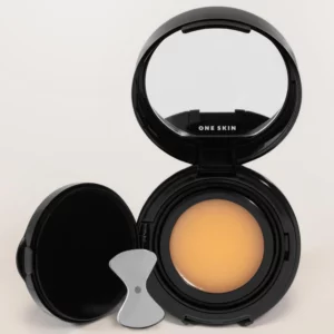 ONE SKIN - kosmetyk do makijażu twarzy w ciepłym beżowym odcieniu, przedstawiony w otwartym, eleganckim czarnym kompakcie z lusterkiem, wewnętrzną pokrywką ochronną i aplikatorem. Minimalistyczne opakowanie na jasnym, neutralnym tle.