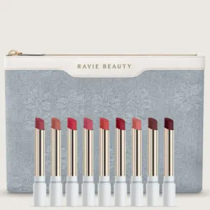 Start 52 Dziewięć odcieni pomadki Ravie Beauty Effortless Lips™ Lip Color ustawionych w rzędzie na tle eleganckiej, błękitnej kosmetyczki z haftowanym wzorem i logo Ravie Beauty. Pomadki w jasnoniebieskich opakowaniach ze złotymi detalami prezentują gamę kolorów od delikatnych różów i brzoskwiniowych nude po intensywne czerwienie, brązy i odcienie jagodowe.